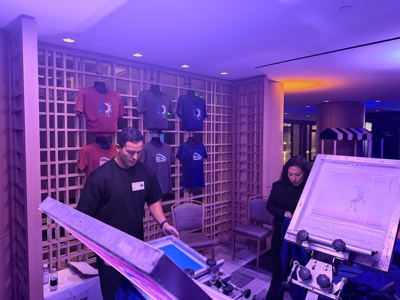 Two printers working a screen-printing station in front of a lattice wall of printed shirts at a purple-lit reception