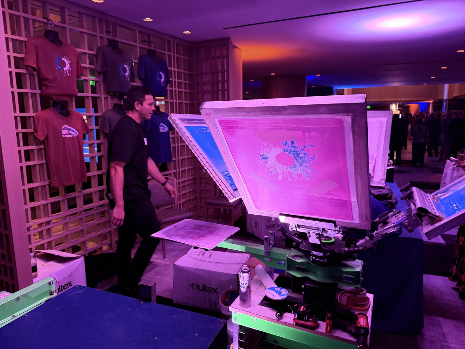 Close-up of a pink-lit screen flooded with ink on a four-color press beside a wall of printed shirts