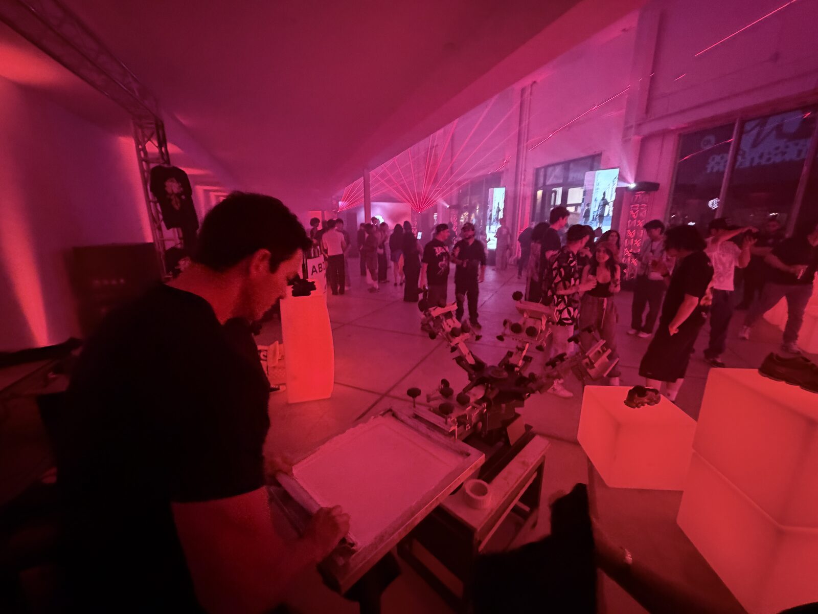 Printer flooding a screen with ink at a four-color press while a crowd mingles at a red-lit night party
